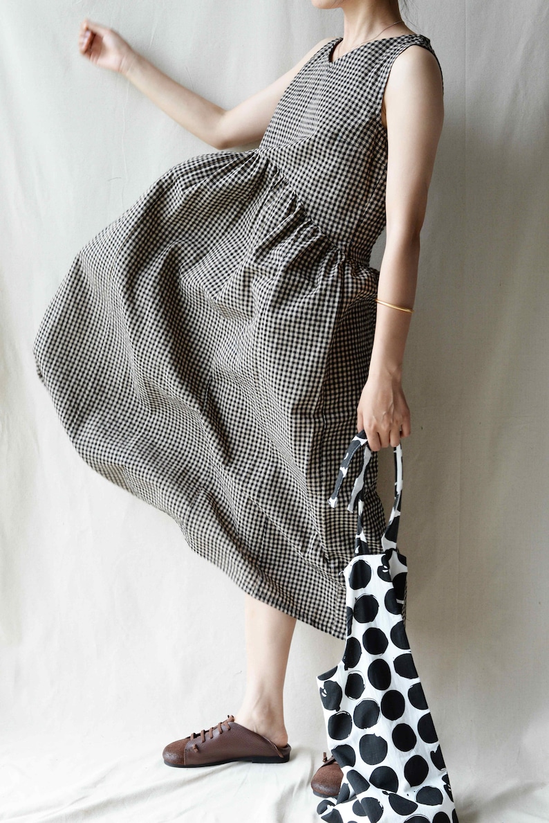 May include: A black and white gingham dress with a loose, flowing silhouette. The dress has a sleeveless design and a round neckline. The model is wearing brown leather shoes.
