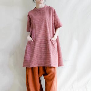 Può includere: Una tunica a quadri rossi e bianchi con maniche corte e due tasche frontali. La tunica è abbinata a pantaloni larghi color ruggine. L'outfit ha uno stile rilassato e casual.