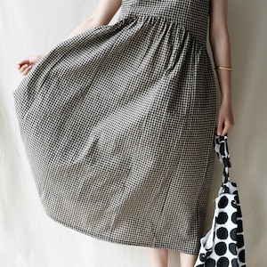 May include: A sleeveless, black and white gingham dress with a gathered waistline. The dress is midi length and has a v-neckline. The model is wearing brown shoes and holding a black and white polka dot tote bag.