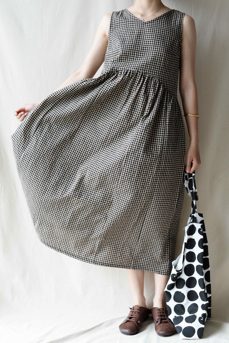 May include: A sleeveless, black and white gingham dress with a gathered waistline. The dress is midi length and has a v-neckline. The model is wearing brown shoes and holding a black and white polka dot tote bag.