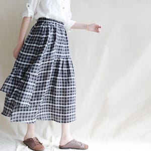 Può includere: Una gonna midi a quadri bianchi e neri con un design a balze. La gonna è abbinata a una camicetta bianca. Il motivo della gonna è un classico motivo a quadretti gingham, che offre un'estetica senza tempo. La gonna è realizzata in tessuto leggero.