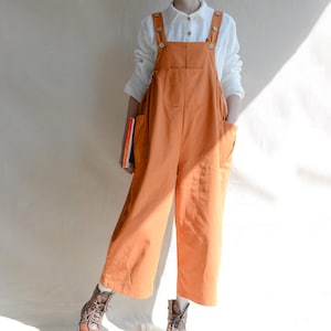 May include: An orange jumpsuit with wide legs and large front pockets. The jumpsuit has button closures on the straps and is worn over a white button-down shirt. The model is wearing brown boots and holding a book.