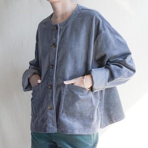 May include: A slate blue corduroy jacket with a round collar and large patch pockets. The jacket has a button-up front and long sleeves with rolled cuffs. The garment is made of a ribbed fabric and has a relaxed fit.