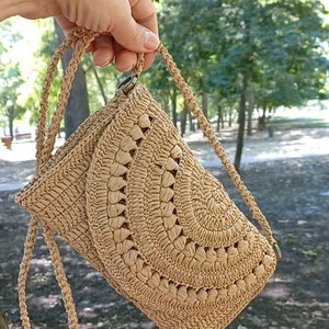 Puede incluir: Un bolso de hombro de crochet de color beige con un patr&oacute;n circular. El bolso tiene una correa larga y un peque&ntilde;o cierre.