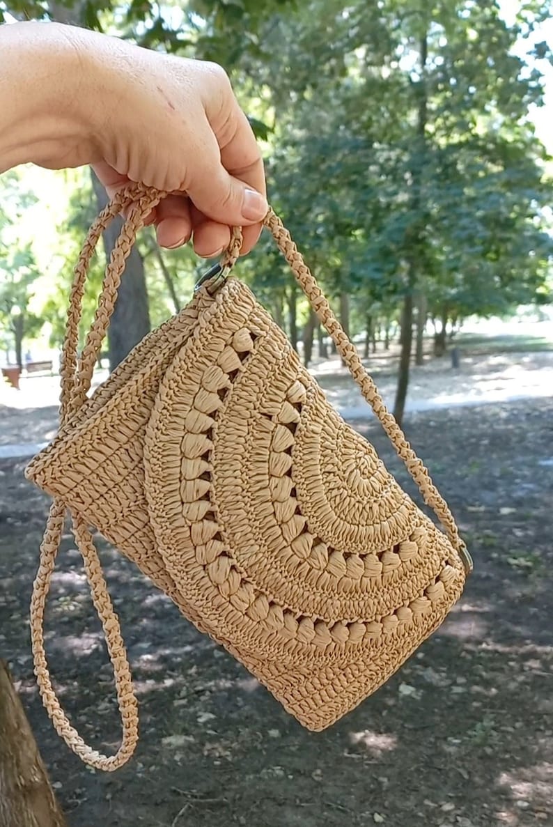Puede incluir: Un bolso de hombro de crochet de color beige con un patr&oacute;n circular. El bolso tiene una correa larga y un peque&ntilde;o cierre.