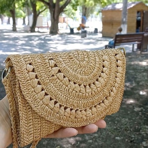 Puede incluir: Un bolso de crochet de color beige con cierre de solapa y una correa larga. El bolso tiene un dise&ntilde;o tejido con recortes circulares.
