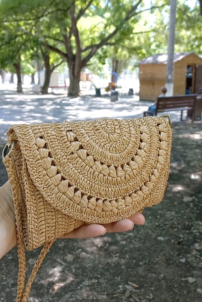 Puede incluir: Un bolso de crochet de color beige con cierre de solapa y una correa larga. El bolso tiene un dise&ntilde;o tejido con recortes circulares.