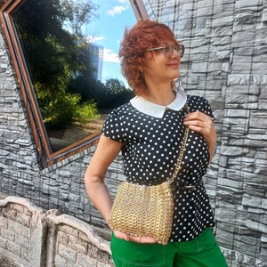 May include: A small, gold-coloured shoulder bag with a chain strap. The bag is made of interlocking circular pieces. The person in the image is wearing a black and white polka dot top, green trousers, and glasses.