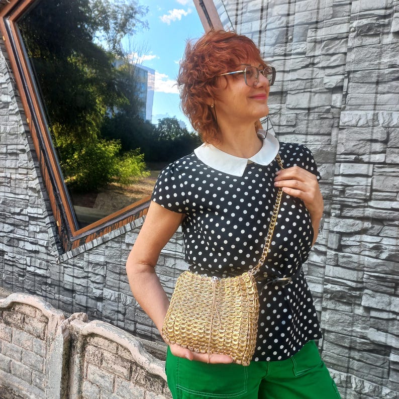 May include: A small, gold-coloured shoulder bag with a chain strap. The bag is made of interlocking circular pieces. The person in the image is wearing a black and white polka dot top, green trousers, and glasses.