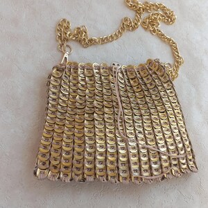 May include: A gold-coloured shoulder bag, handmade from numerous metallic, circular pieces, creating a textured surface. It has a gold chain strap and a drawstring closure. The bag's design is unique and eye-catching, a distinctive accessory.