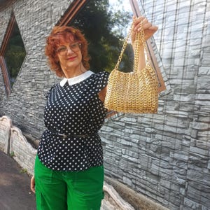 May include: A gold-coloured handbag with a chain strap. The bag is made of interlocking metal pieces. The person in the image is wearing a black and white polka dot top, green trousers, and glasses.