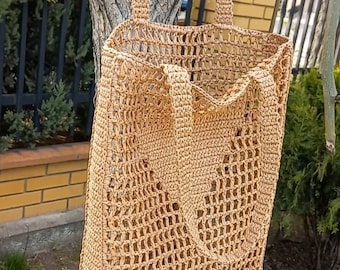 May include: A tan crocheted tote bag with a mesh design. The bag is hanging from a tree branch.