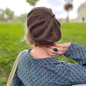 May include: A brown beret with a textured knit pattern. The beret is worn by a person with short brown hair. The person is wearing a blue and white patterned blouse with long sleeves.