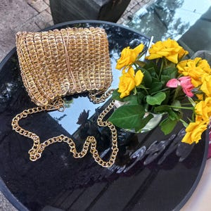 May include: A gold-coloured handbag with a chain strap rests on a black surface beside a vase of yellow and pink roses. The bag seems to be made from woven metal rings, and the chain is a matching gold colour.