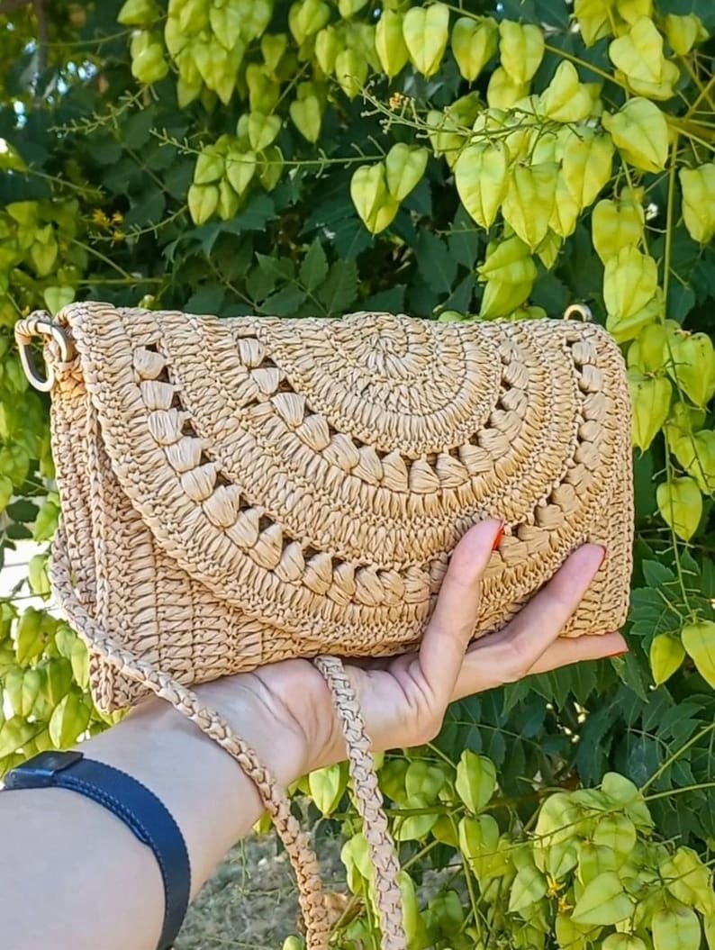 Puede incluir: Un bolso de paja tejido beige con un dise&ntilde;o circular y una correa larga. El bolso est&aacute; hecho de materiales naturales y tiene un estilo informal y veraniego.
