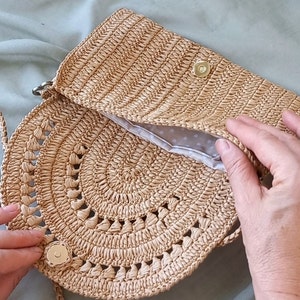 Puede incluir: Un bolso de paja tejido a crochet de color marr&oacute;n claro con una parte superior redonda abierta y un cierre de solapa. El bolso tiene un forro de lunares blancos y un cierre de metal dorado.
