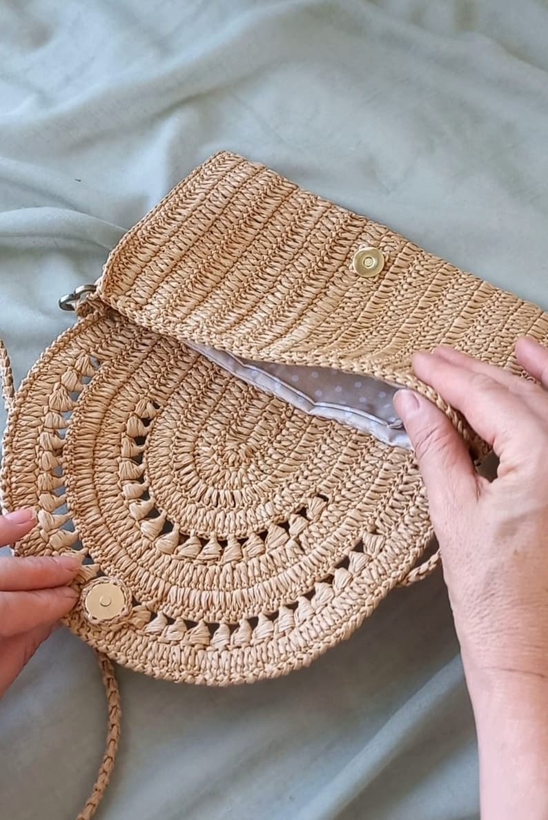Puede incluir: Un bolso de paja tejido a crochet de color marr&oacute;n claro con una parte superior redonda abierta y un cierre de solapa. El bolso tiene un forro de lunares blancos y un cierre de metal dorado.