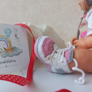 Könnte beinhalten: Ein Paar rosa und weiße gehäkelte Babyschuhe mit grauen Akzenten und weißen Schnürsenkeln. Die Schuhe befinden sich auf einer weißen Oberfläche.