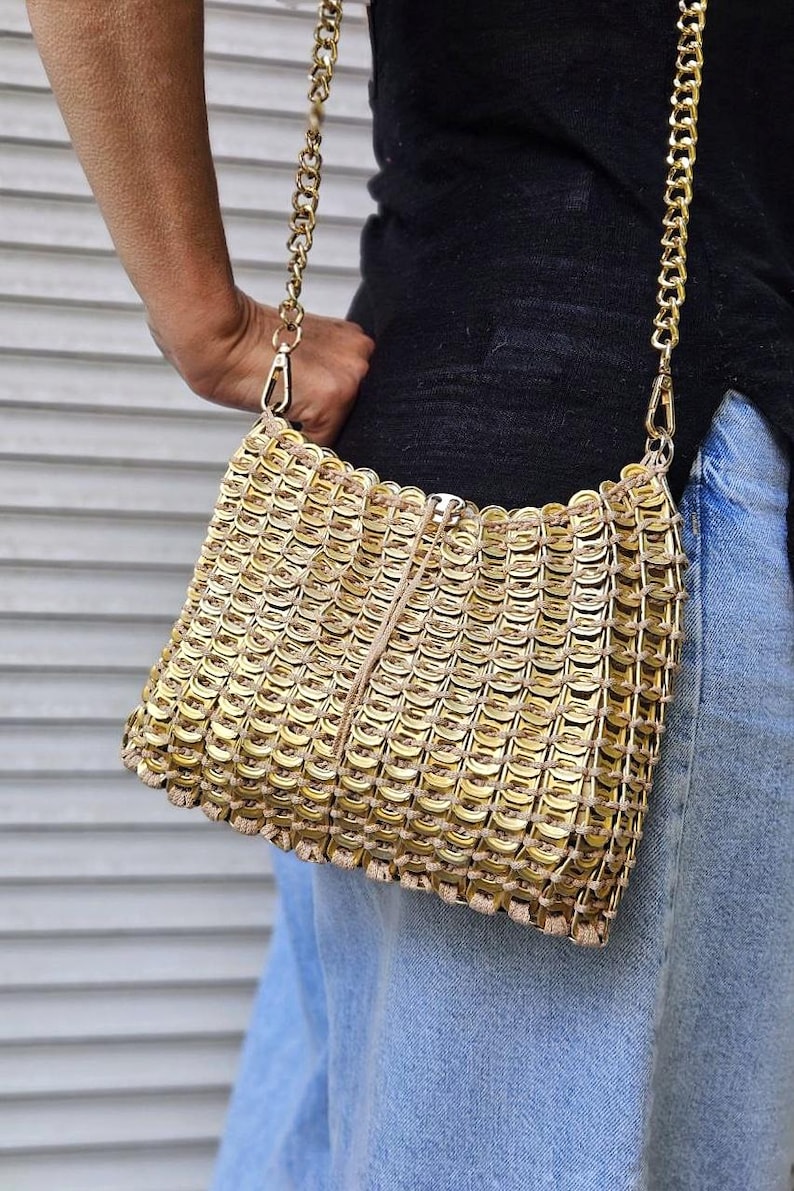 May include: A gold-coloured, rectangular handbag made from recycled can tabs. The bag has a chain strap and a zip closure. The bag is held by a person wearing a black top and blue jeans.