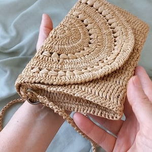 Puede incluir: Un bolso de paja tejido de color marr&oacute;n claro con una solapa redondeada y una correa larga. El bolso tiene un patr&oacute;n circular.