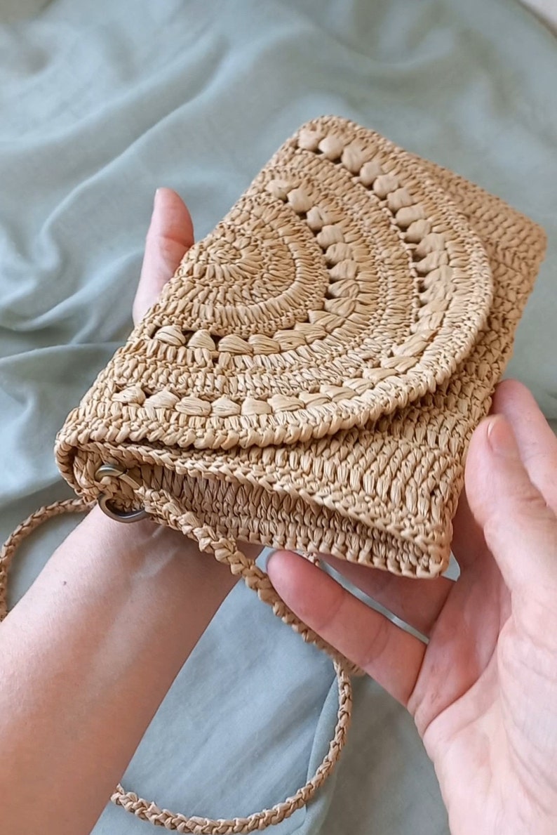 Puede incluir: Un bolso de paja tejido de color marr&oacute;n claro con una solapa redondeada y una correa larga. El bolso tiene un patr&oacute;n circular.
