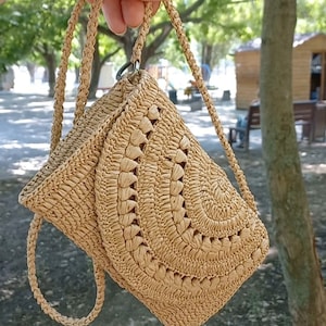 Puede incluir: Un bolso de mano de color beige tejido a crochet con un dise&ntilde;o circular y una correa larga.
