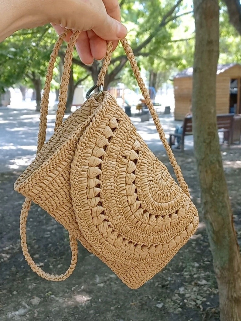 Puede incluir: Un bolso de mano de color beige tejido a crochet con un dise&ntilde;o circular y una correa larga.