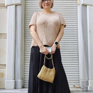 May include: A woman in a beige knit top and a long black pleated skirt, holding a gold chain bag and a white phone. She wears black flats and a watch. The background shows a white shutter door. The outfit is suitable for a casual occasion.