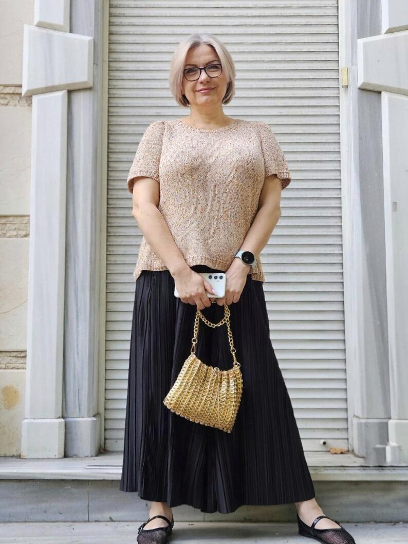 May include: A woman in a beige knit top and a long black pleated skirt, holding a gold chain bag and a white phone. She wears black flats and a watch. The background shows a white shutter door. The outfit is suitable for a casual occasion.