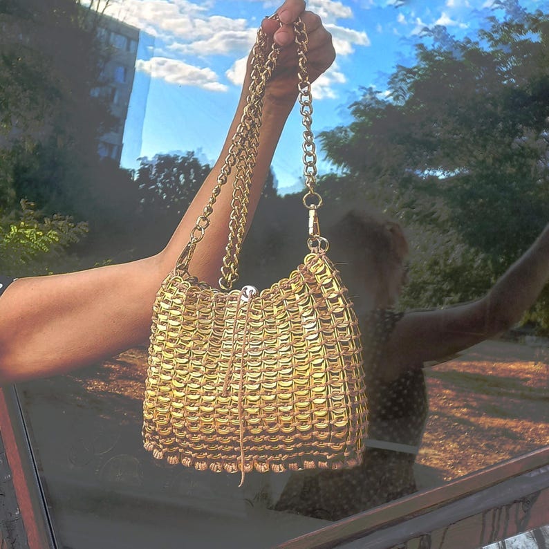 May include: A gold-coloured handbag with a chain strap. The bag is made of interlocking metal pieces, creating a textured surface. The chain strap is also gold and attached to the bag with a clasp.