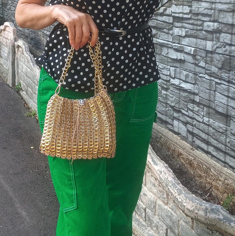 May include: A gold-coloured handbag made from recycled can tabs, with a chain strap. The bag has a rectangular shape with a rounded bottom. The person is wearing a black and white polka dot top and green trousers.
