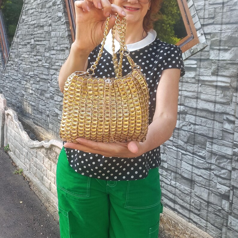 May include: A gold-coloured handbag with a chain strap. The bag is made from interlocking metal pieces, creating a textured surface. The bag is held up in front of a grey stone wall.