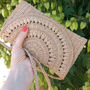 Puede incluir: Un bolso de crochet beige con una correa larga. El bolso tiene un patr&oacute;n circular y est&aacute; hecho de fibras naturales.