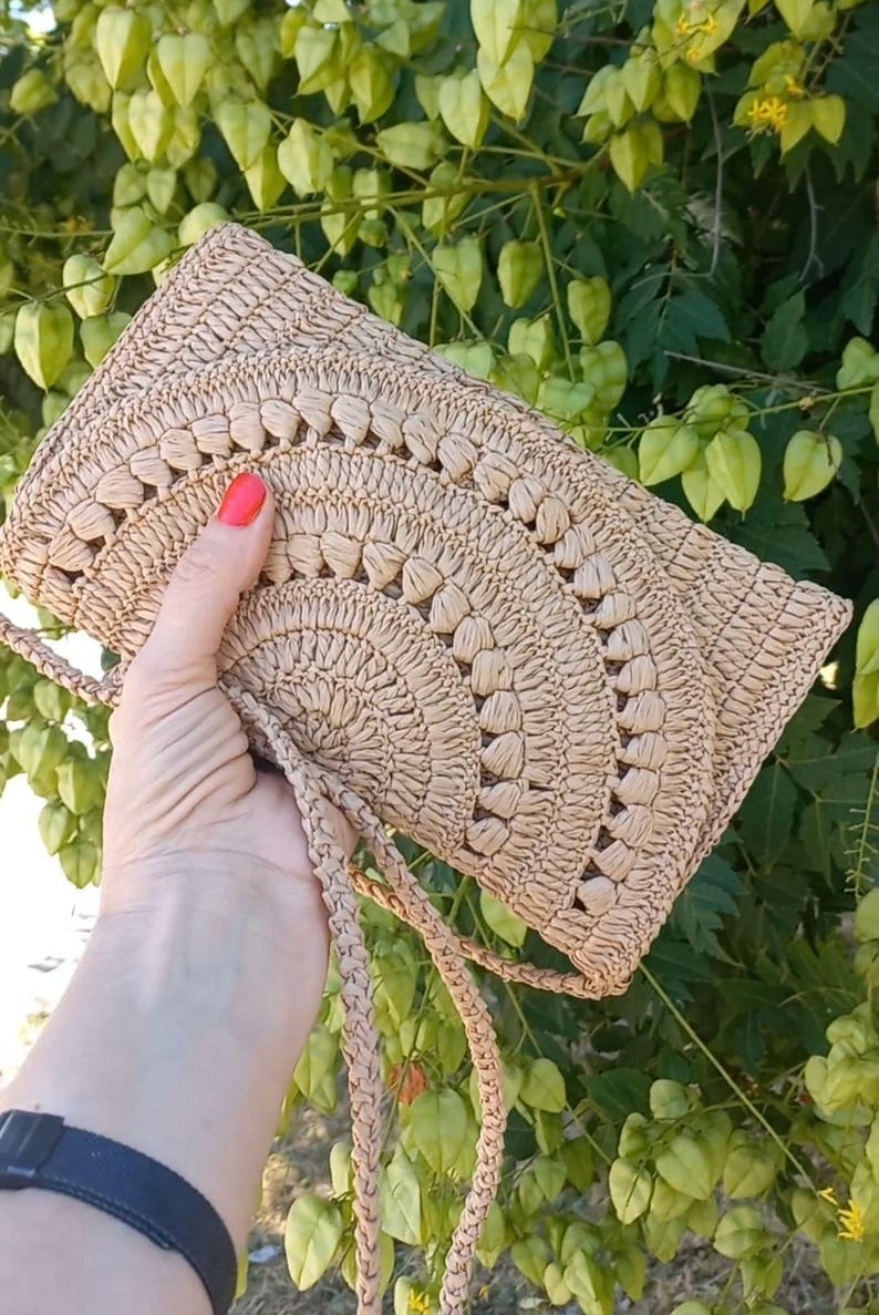 Puede incluir: Un bolso de crochet beige con una correa larga. El bolso tiene un patr&oacute;n circular y est&aacute; hecho de fibras naturales.