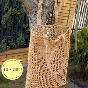 Op de afbeelding: Een bruin gehaakte tote bag met een rechthoekige vorm en open weefsel. De tas heeft twee handvatten en hangt aan een tak. De tekst "PDF + VIDEO" staat in een gele cirkel en "CROCHET PATTERN" staat eronder.
