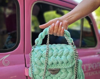 May include: A light green crocheted handbag with a silver chain strap and a white tassel. The bag has a braided design and white polka dots.