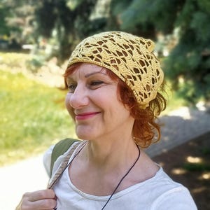 Pode incluir: Um chapéu de crochê amarelo claro com um design rendado. A pessoa que o usa tem cabelo ruivo e usa uma camiseta branca com gráficos de gatos pretos. O fundo é uma cena externa desfocada com folhagem verde.