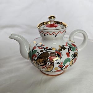 Könnte beinhalten: Eine weiße Porzellan-Teekanne mit goldener Verzierung und einem Hahn-Design. Die Teekanne hat einen roten und goldenen Deckel und einen roten und goldenen Griff.