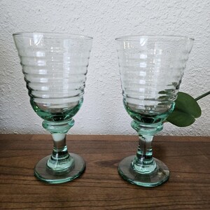 Puede incluir: Dos copas de vino de vidrio transparente con un tinte verde y un diseño acanalado. Las copas tienen un tallo y una base redonda.