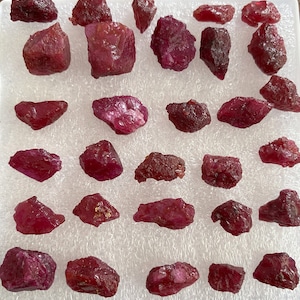 May include: A collection of raw, uncut ruby gemstones. The rubies vary in size and shape, displaying a deep red hue with some variations in tone. They are arranged on a white, textured surface, showcasing their natural, unpolished state.
