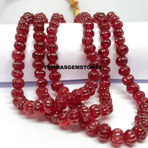 May include: A strand of deep red ruby beads, each bead is carved with a textured pumpkin shape. The beads are strung together on a clear thread and are displayed on a white surface.  The text "THOMASGEMSTONES" is visible in the image.