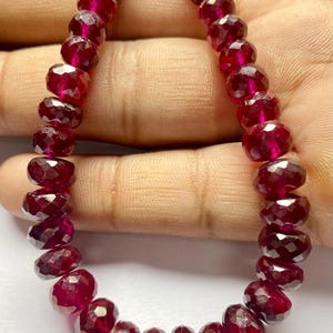 Puede incluir: Un collar de cuentas de piedras preciosas facetadas de color rojo intenso. Las cuentas tienen forma ovalada y están ensartadas, mostrando un color rico y translúcido. Las piedras preciosas parecen pulidas, reflejando la luz y añadiendo un toque de elegancia.