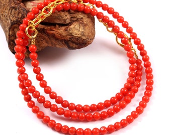 Puede incluir: Un collar de cuentas de coral con un cierre de cadena de color dorado. El collar está compuesto por pequeñas cuentas redondas de color naranja brillante. La cadena dorada añade un toque de elegancia. El collar se exhibe sobre un trozo de madera.
