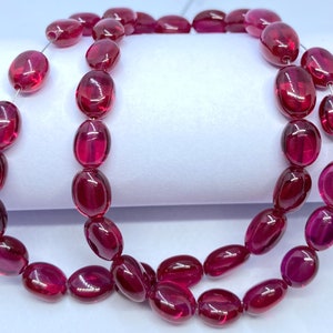 Può includere: Una collana di perle ovali di pietra preziosa di colore rosso scuro. Le perle sono infilate su un filo sottile e sono disposte in un motivo sciolto e fluido.