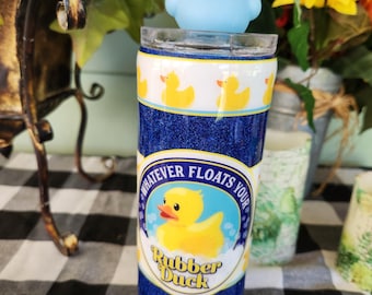 May include: A blue and white tumbler with a glitter finish. The tumbler has a yellow rubber duck design with the text "Whatever Floats Your Rubber Duck". A blue rubber duck is on top of the tumbler.