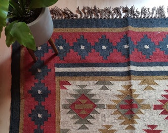 May include: A woven rug with a geometric pattern in shades of red, brown, white, and black. The rug has a fringe border.