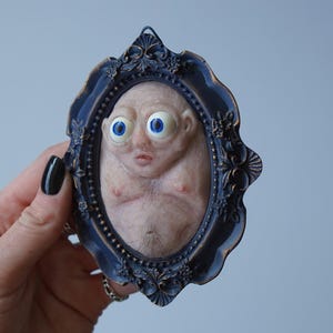 Op de afbeelding: Een ovale sculptuur van een wezen met grote blauwe ogen en een bleke huid, geplaatst in een donkerblauwe, sierlijke lijst. De sculptuur heeft een gedetailleerd, getextureerd uiterlijk, en de lijst heeft een vintage esthetiek.