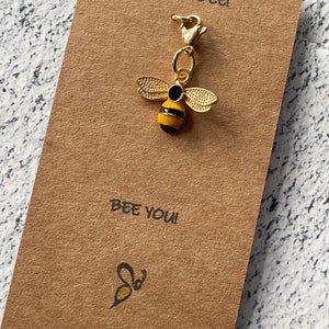 May include: A gold bee charm with black stripes and a gold clasp. The charm is on a brown card with the text "Just Bee!" and "Bee You!"