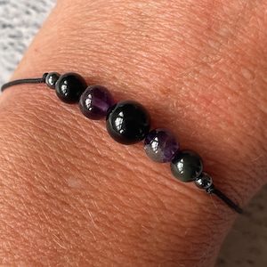 May include: A black string bracelet with black, purple, and silver beads. The bracelet is on a person's wrist.