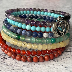 May include: Seven chakra bracelets stacked on top of each other. The bracelets are made of different colored stones, including red, orange, yellow, green, blue, indigo, and violet. A silver charm with the symbol "Om" is attached to the top bracelet.